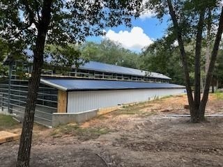 Long white metal barn with a black roof and windows, set in a wooded area.