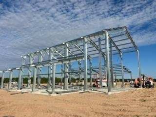 Steel framework of a building under construction on a dirt lot, against a blue sky with clouds.