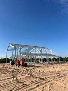 Construction of a steel-framed building under a blue sky, with heavy machinery on a dirt lot.