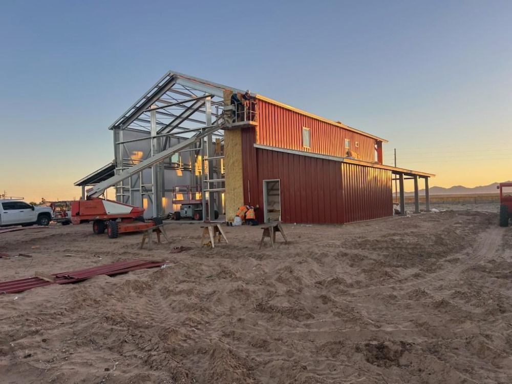 Construction of a red metal barn in a desert setting with workers using a lift.