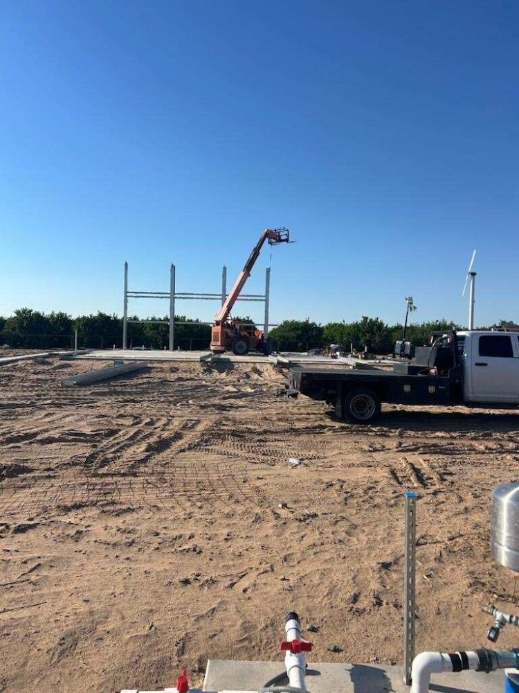 Construction site with steel beams, an orange lift, and a flatbed truck on dirt under a blue sky.