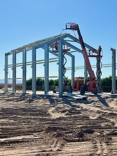Construction of a metal building with workers on lifts, setting beams on a sunny day.