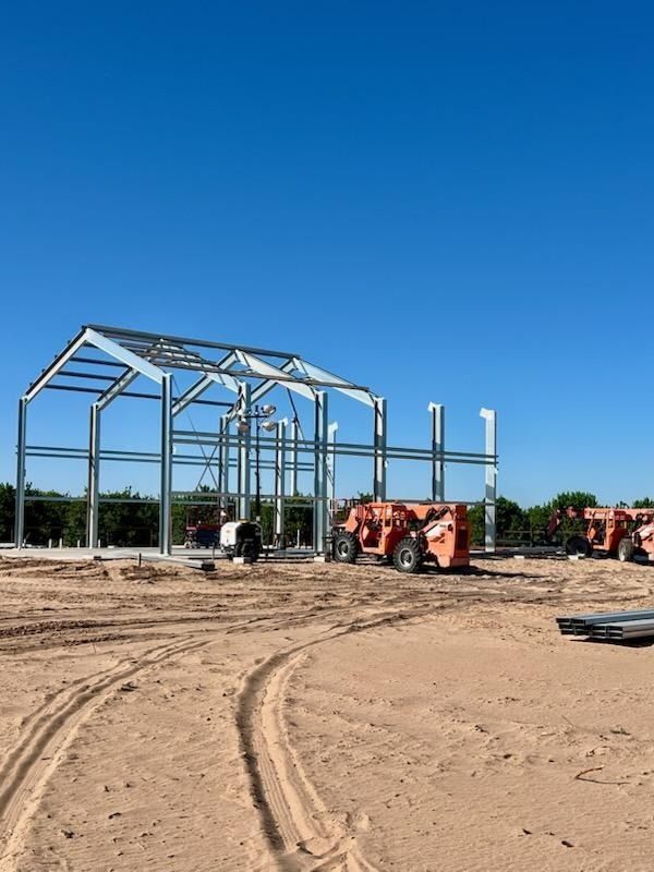 Construction site with steel frame building under construction, orange lift vehicles on dirt, clear blue sky.