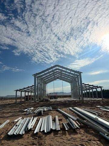 Steel frame of a building under construction on a dirt lot, bright sky overhead with some clouds and the sun.