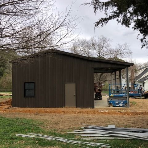Brown metal building with lean-to carport. Door, window, construction materials on ground, trees in the background.