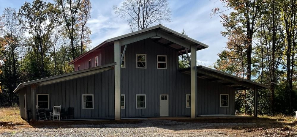 A two-story gray building with a red roof sits amongst trees. It has a covered porch.