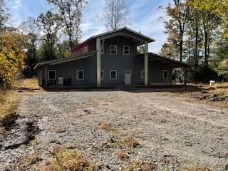 Gray two-story house with a gravel driveway, surrounded by trees under a blue sky.