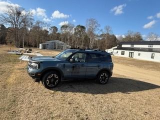 Blue Ford Bronco Sport parked on dry grass with buildings and trees in the background.
