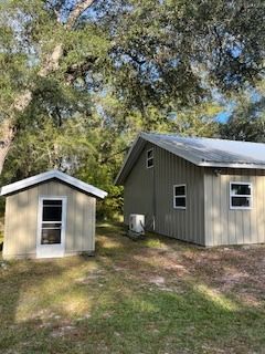 Two beige metal-sided buildings with white-framed windows, set on a grassy lot, shaded by trees.