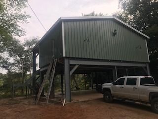 Green metal-sided elevated structure with a pickup truck parked underneath and a wooden ladder leaning against it.