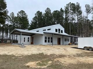 White metal-sided building with a covered porch and a trailer in a wooded area.