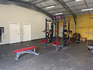 Gym interior with weight rack, bench, and equipment on a dark floor. Two white doors on the left.