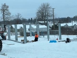 Construction site in winter with steel frame building under assembly. A worker in orange vest is present. Snowy ground.