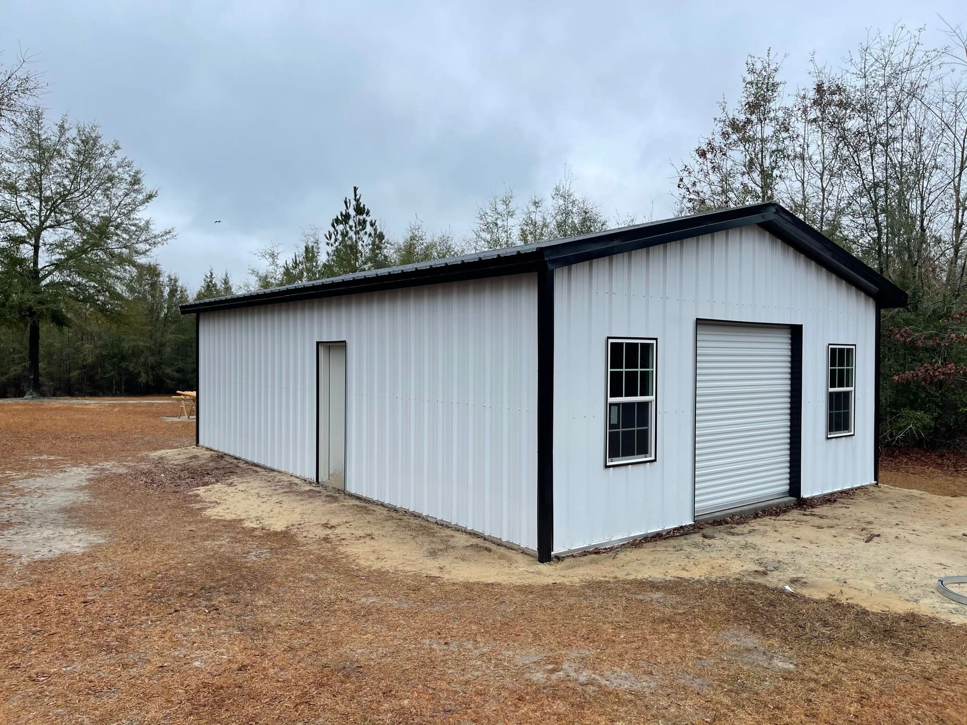 White metal building with black trim, a garage door, and windows on a gravel lot, surrounded by trees under a cloudy sky