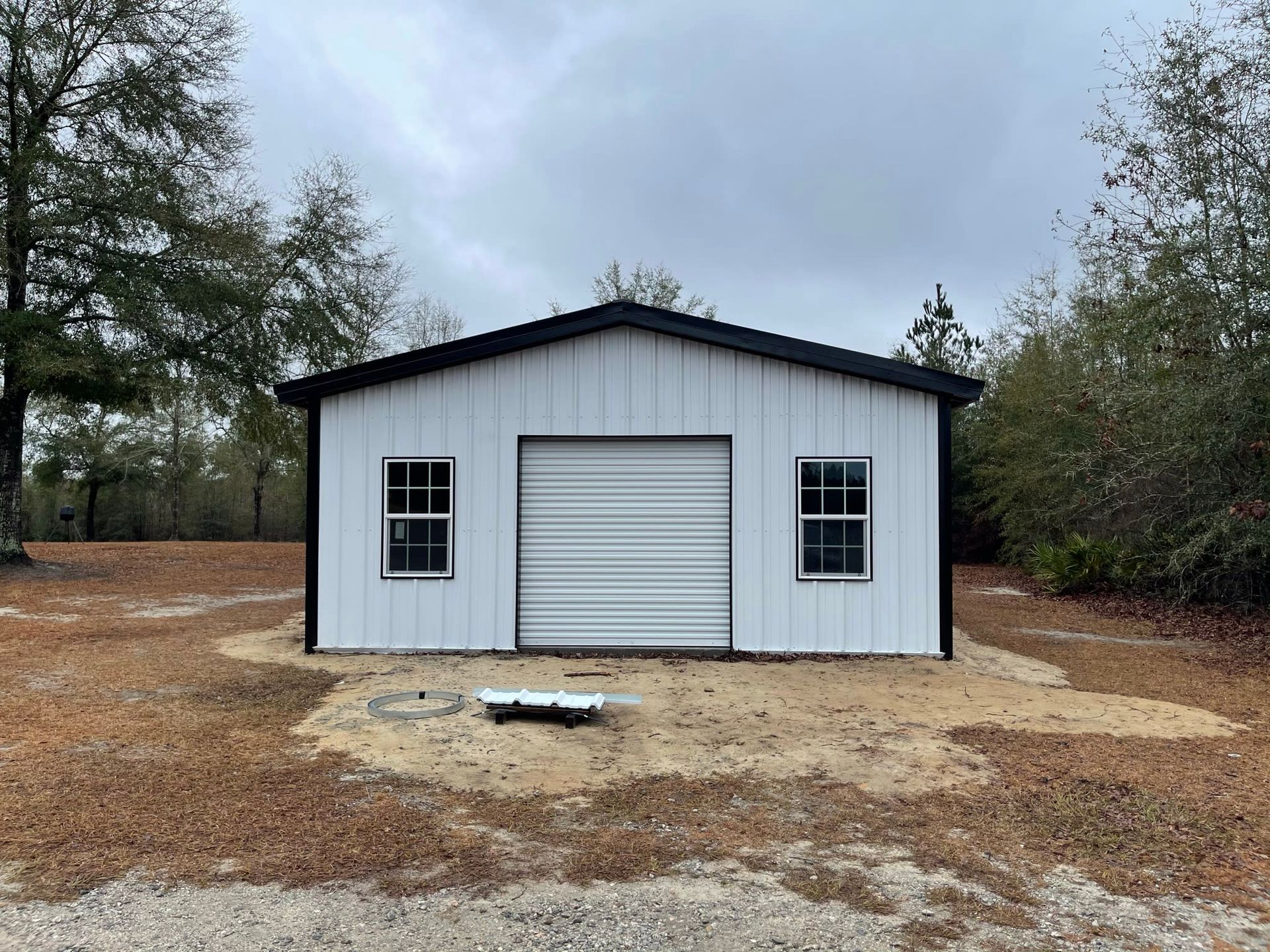 White metal garage with black trim and garage door, two windows, on a sandy lot under cloudy sky