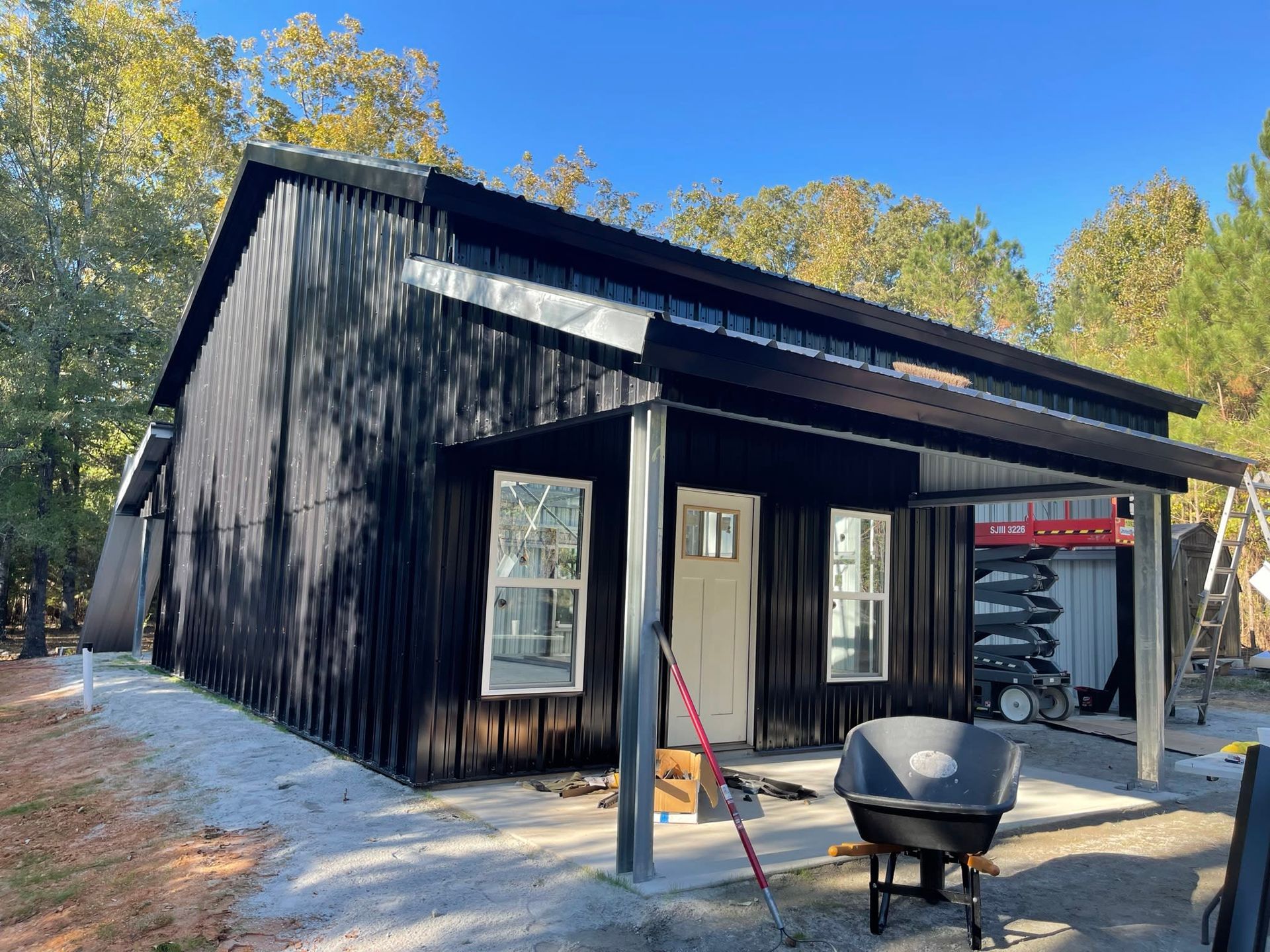 Black metal-sided building with a covered porch, windows, and a white door, set in a wooded area