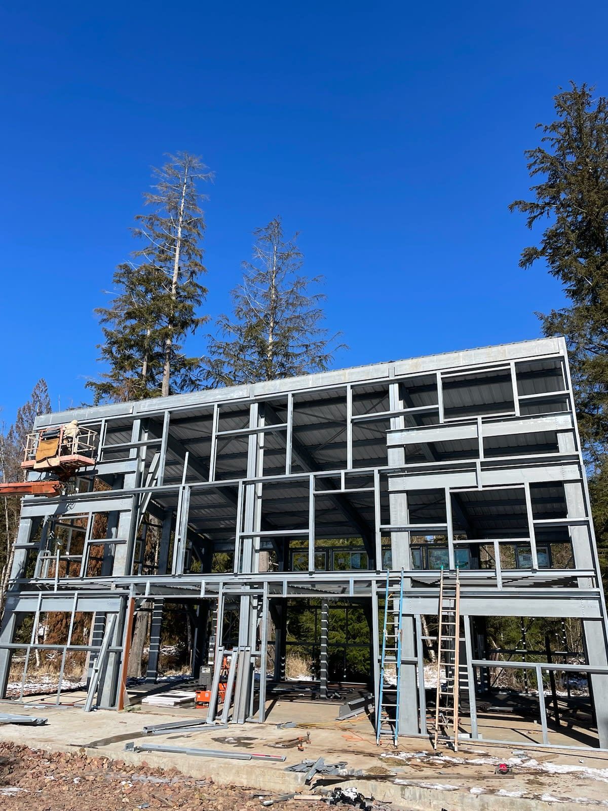 Steel frame of a two-story house under construction against a blue sky, trees in the background