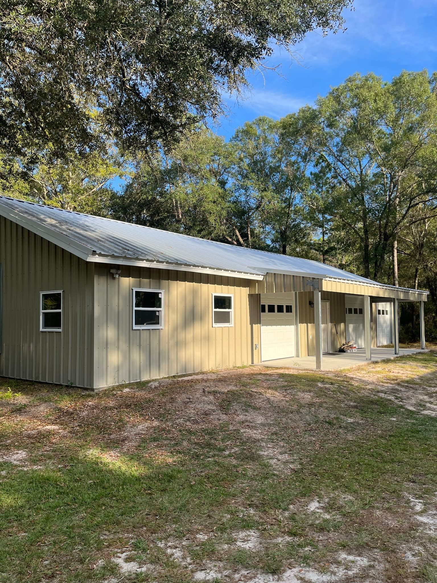 Tan building with a corrugated metal roof, two windows, and a garage door, set among trees under a blue sky
