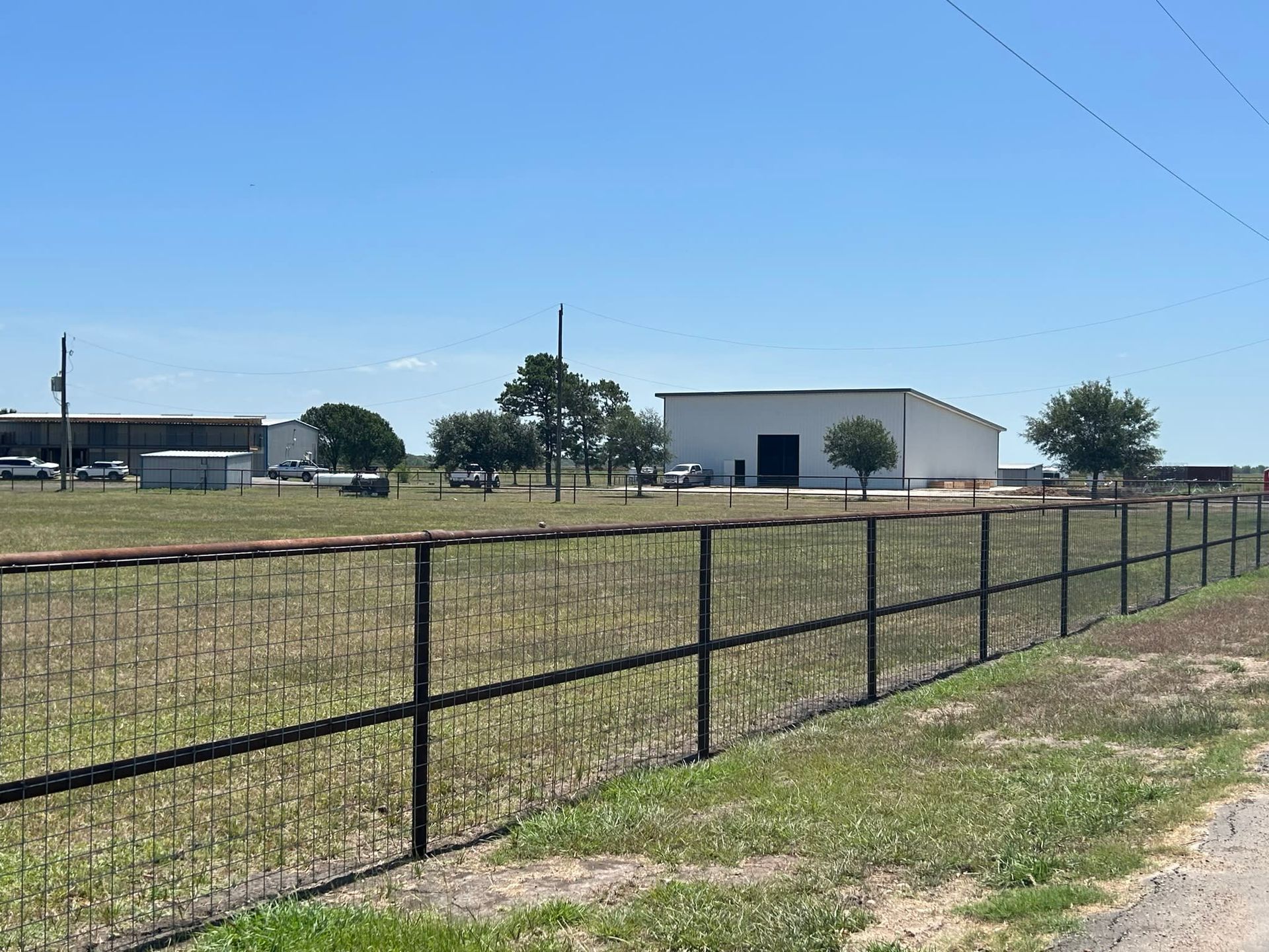 A black metal fence borders a grassy field with industrial buildings under a blue sky
