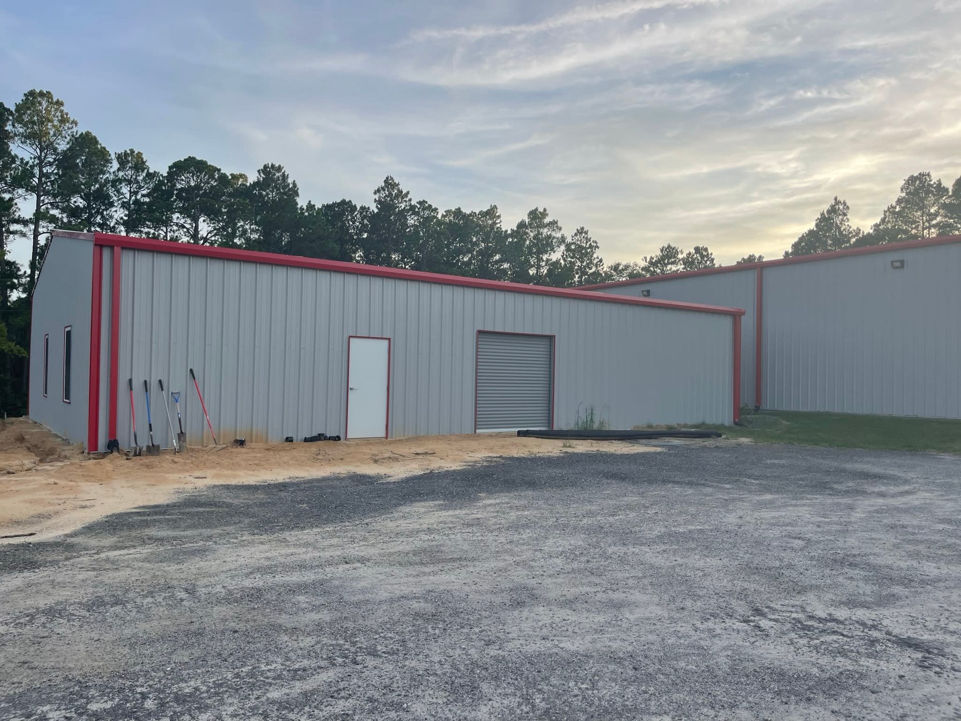 Gray metal building with red trim, gravel lot, cloudy sky, and trees