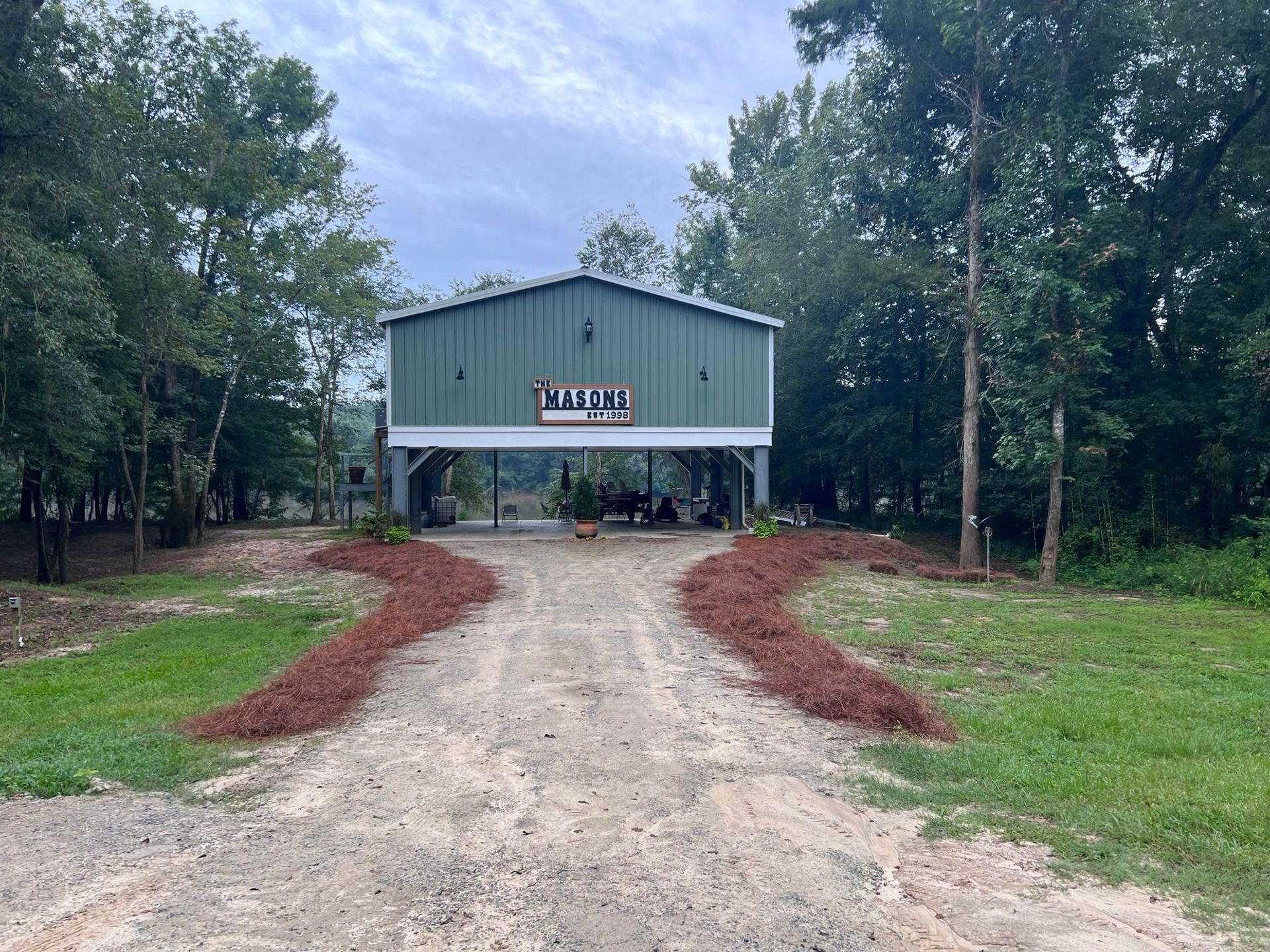 Driveway leads to a green metal building with a sign, surrounded by trees