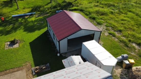 Aerial view of a new red-roofed metal building with an open garage door, surrounded by grass