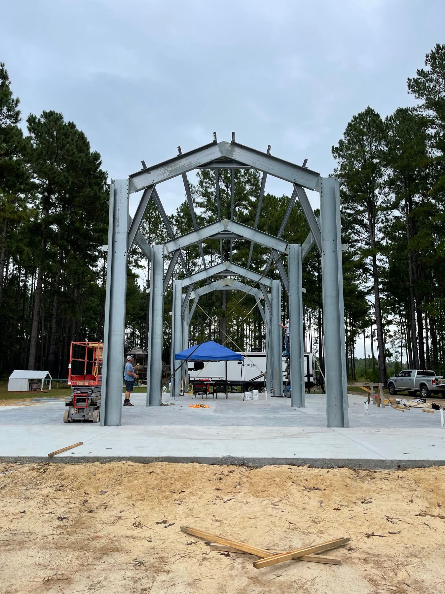 Steel frame of a building under construction on a concrete slab, with workers, in a wooded area