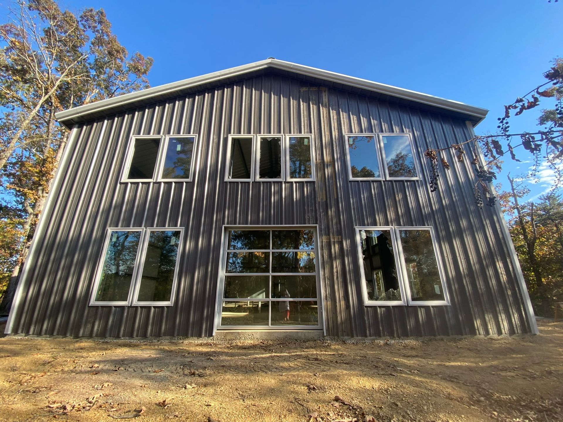Dark gray metal-clad two-story building with multiple windows and a glass door in a wooded area on a sunny day