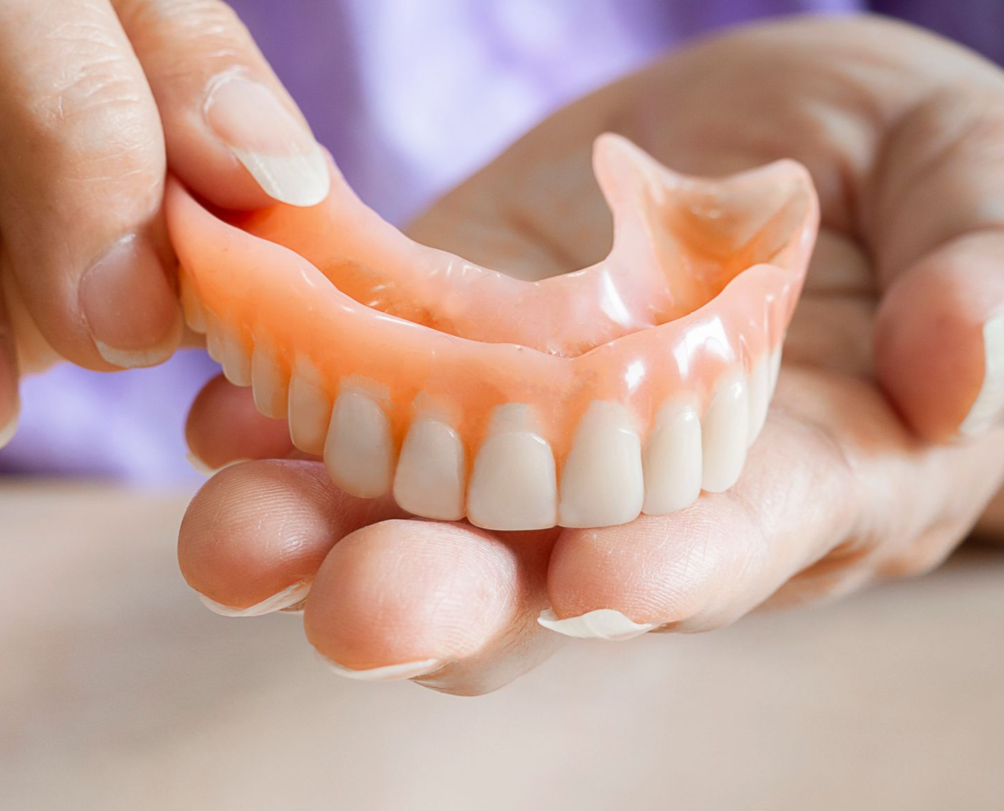Manos sosteniendo una dentadura postiza superior rosa con dientes blancos sobre un fondo borroso.
