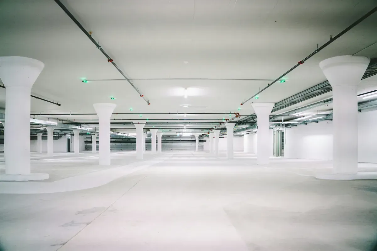 Empty, bright white parking garage with numerous pillars and overhead pipes and lights.