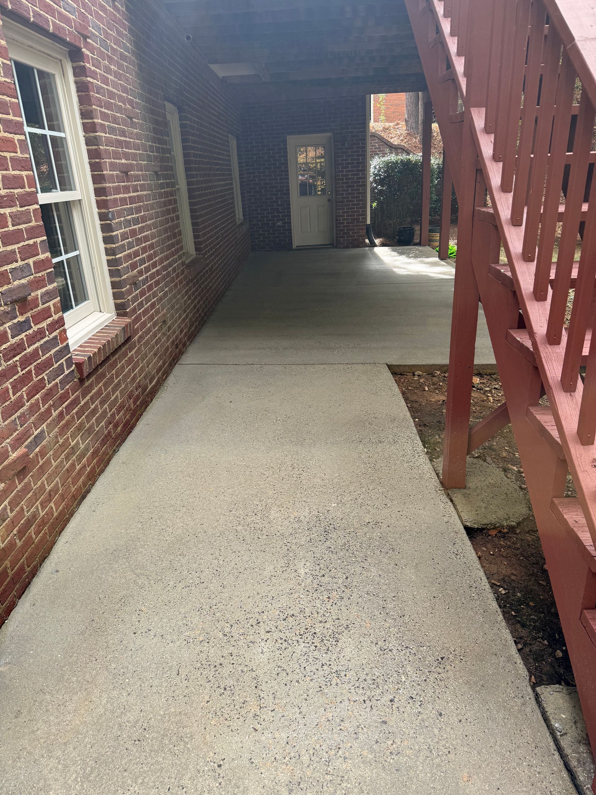 Concrete walkway beside a brick building and wooden stairs, leading to a door under a covered area.