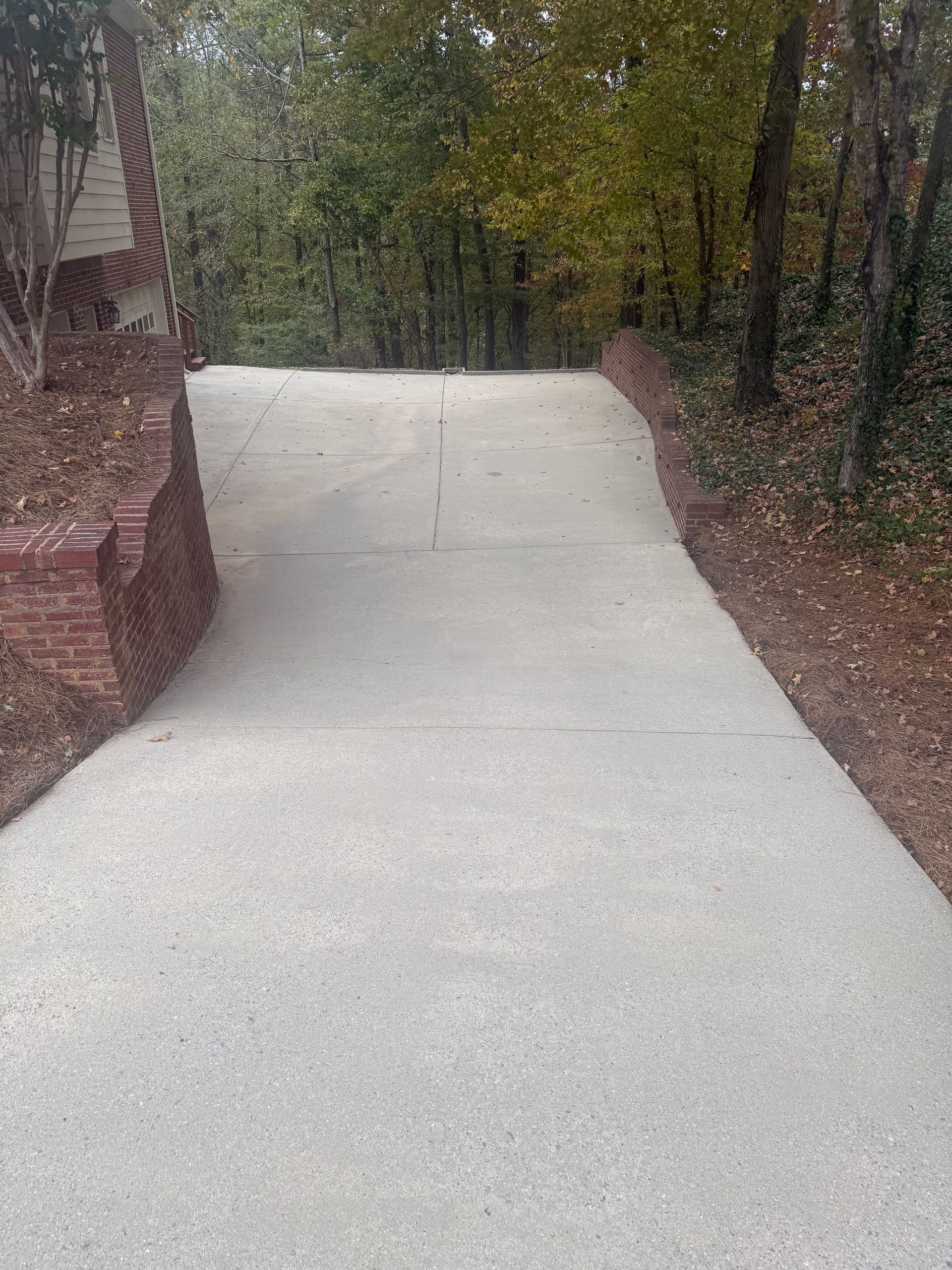 Concrete driveway leading uphill, flanked by brick wall and foliage.