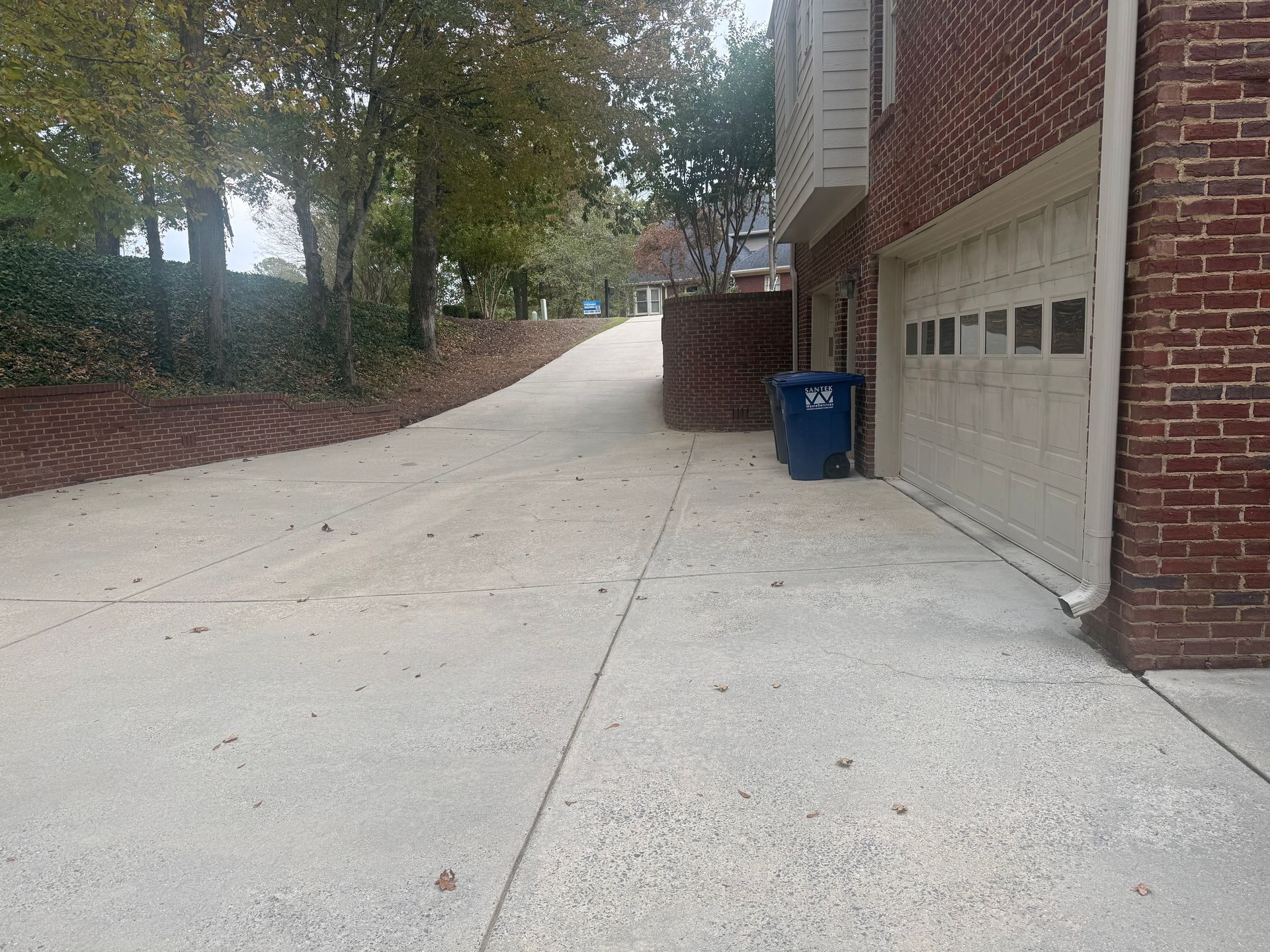 Concrete driveway leading uphill, beside a brick building with a garage door and a blue trash bin.