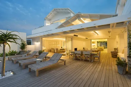 White house with deck, lounge chairs, dining table, and plants; evening setting.