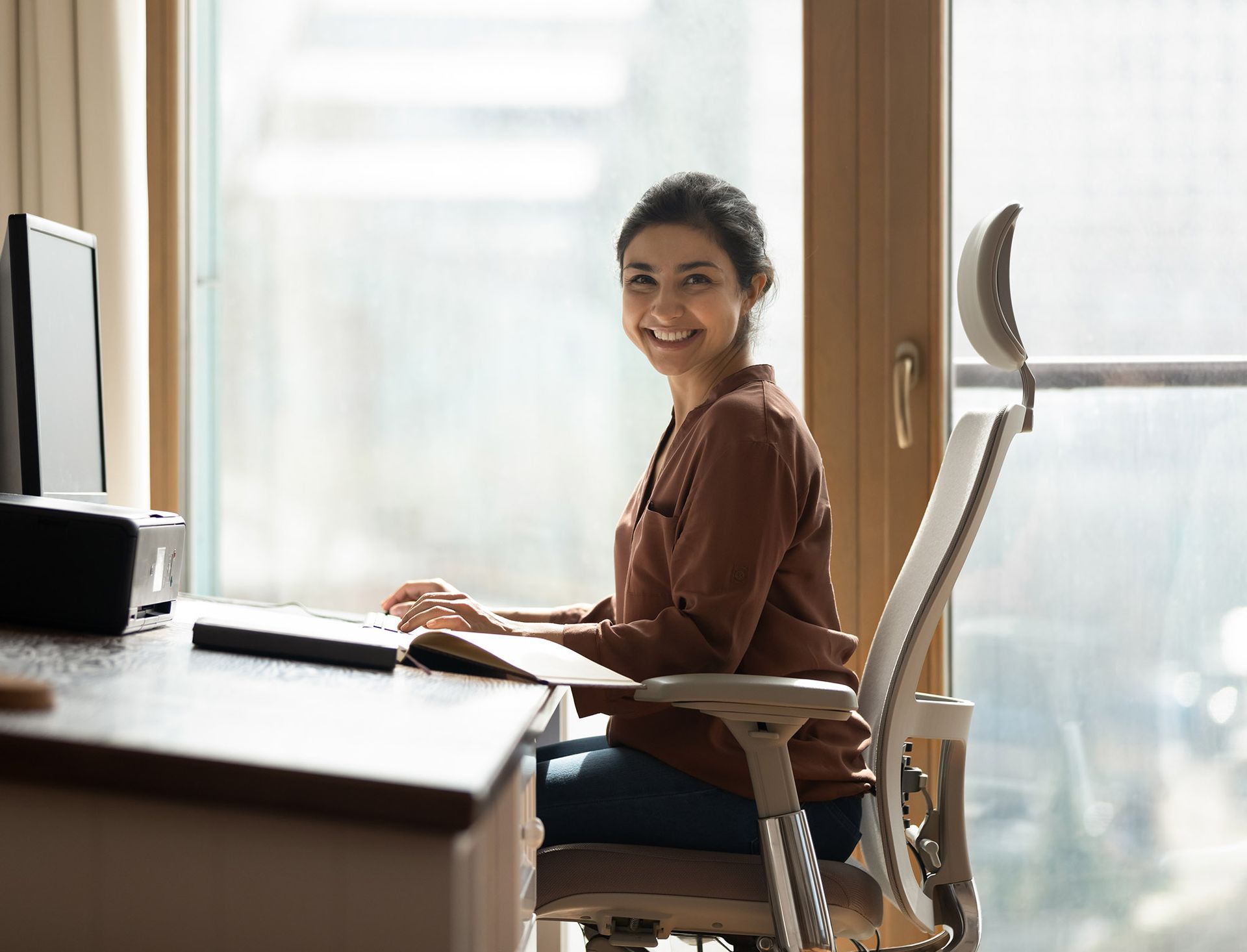 Woman smiles while seated at desk by window, working on computer.