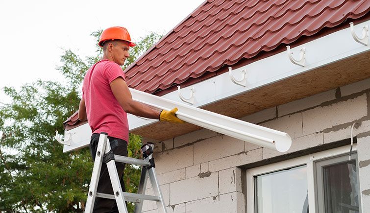 A man is standing on a ladder installing a gutter on the side of a house.