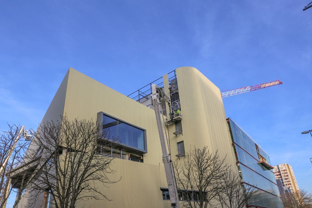 Bâtiment moderne à la façade beige incurvée et aux panneaux de verre ; grue dans le ciel bleu.