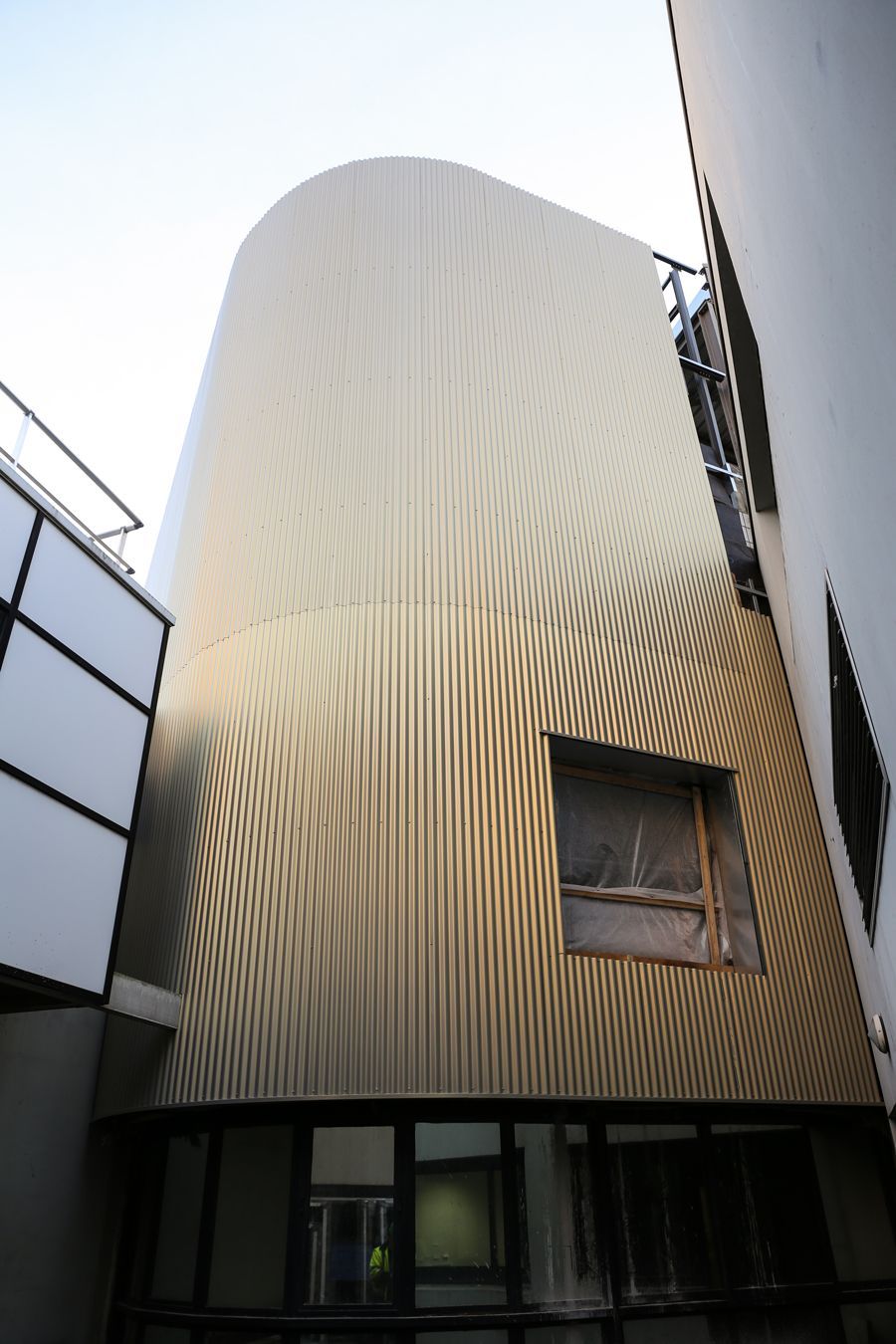 Façade dorée et incurvée d'un bâtiment aux arêtes verticales, entre deux bâtiments blancs, ciel nuageux.