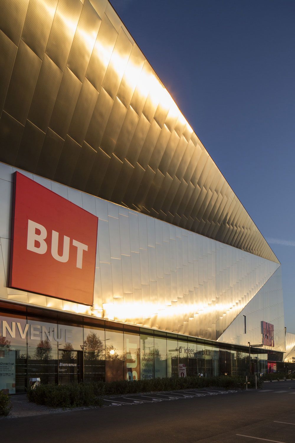 MAIS l'extérieur du magasin, avec sa façade en métal doré et son logo rouge, reflète la lumière.