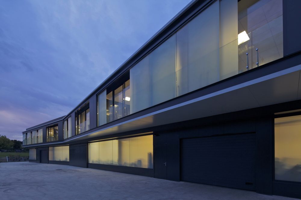 Bâtiment moderne gris foncé, avec de grandes fenêtres dépolies reflétant la lumière intérieure sur un ciel crépusculaire.