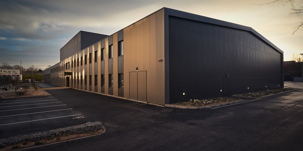 Bâtiment industriel moderne gris foncé, à la façade longue et linéaire. Allée asphaltée et ciel couvert.