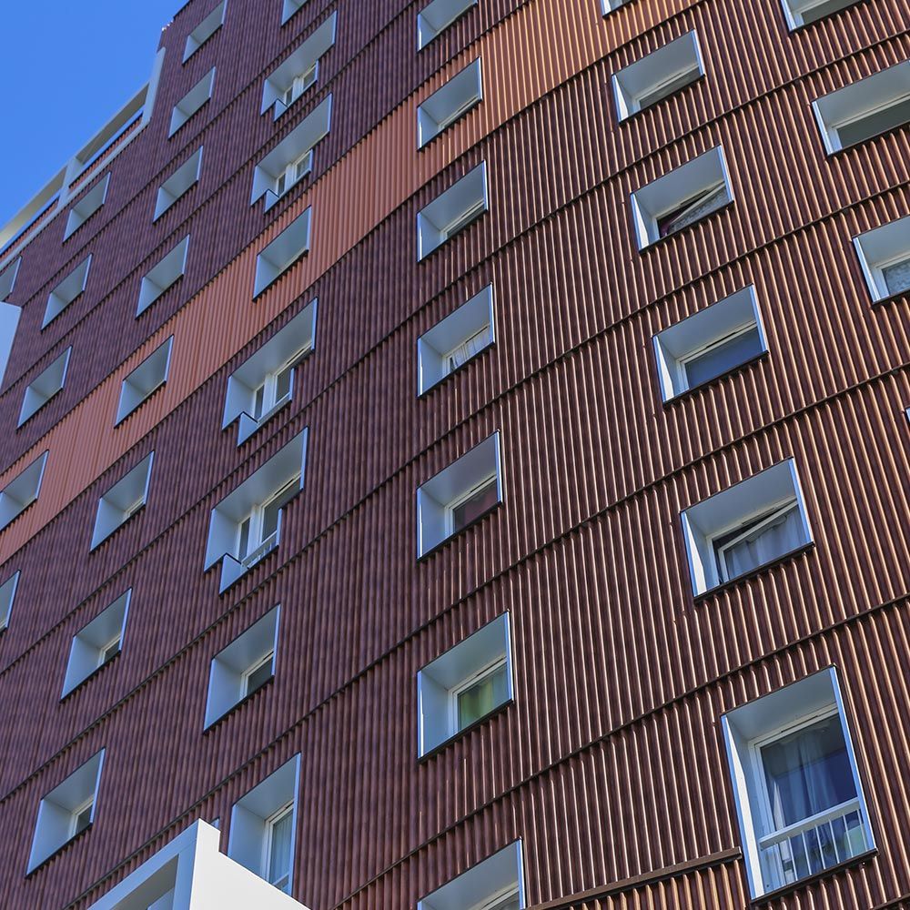 Extérieur du bâtiment avec de nombreuses fenêtres rectangulaires, une façade nervurée brune et un ciel bleu.