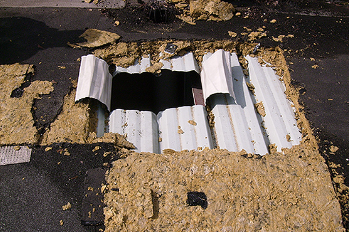 Toiture endommagée présentant un trou rectangulaire, laissant apparaître l'isolant et des éléments de structure.