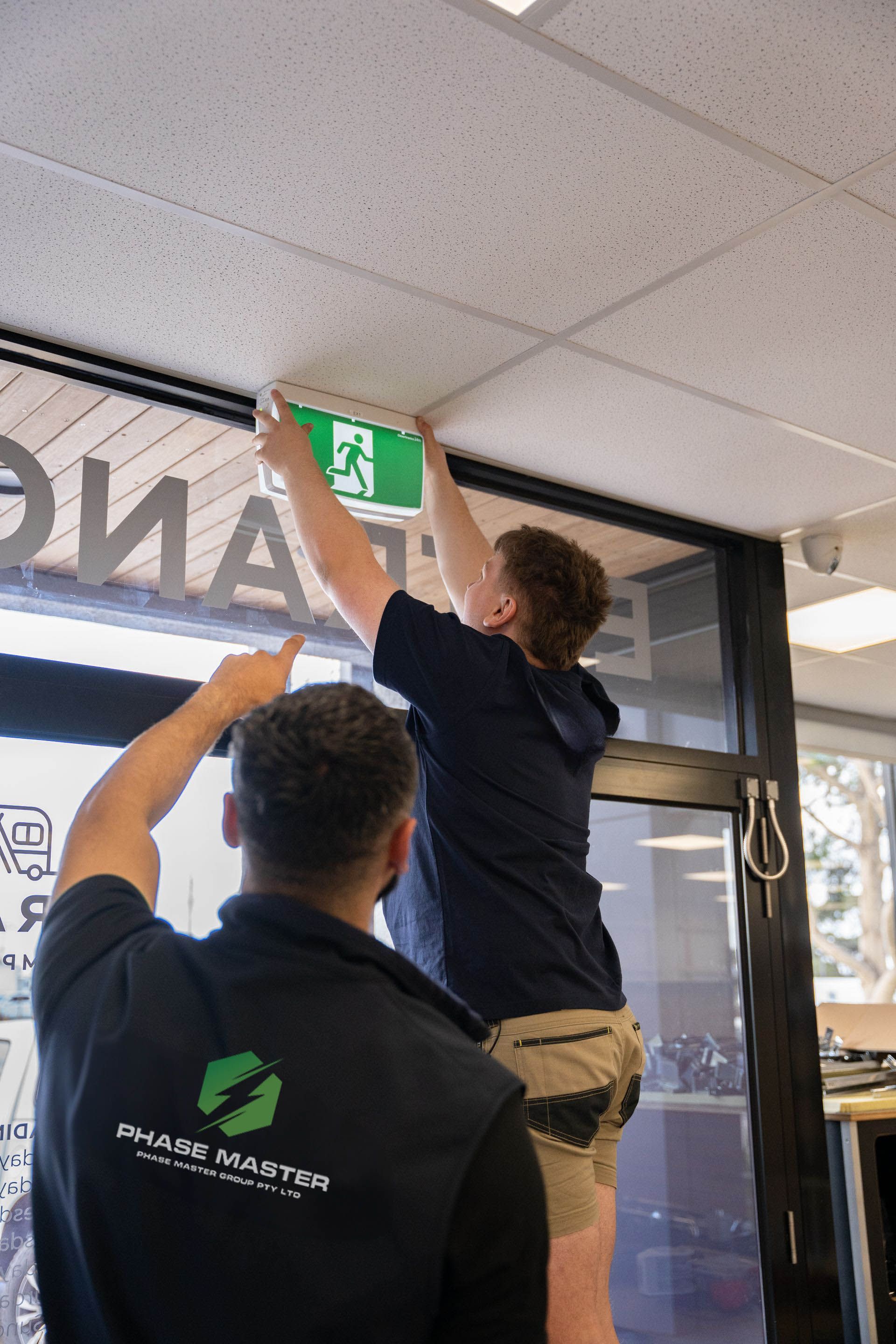 Two men are working on an emergency exit sign in a building.