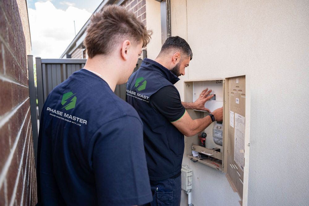 Two men are working on an electrical box on the side of a building.
