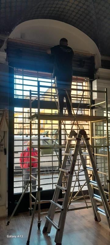 Persona su un'impalcatura che lavora su una finestra con barre di metallo. Un'altra persona è in piedi accanto. Ambientazione interna.