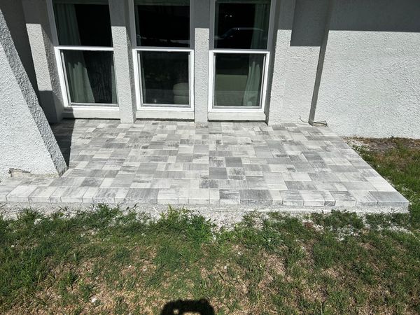 A gray and white stone paver patio situated outside a home with three glass sliding doors and textured light walls.
