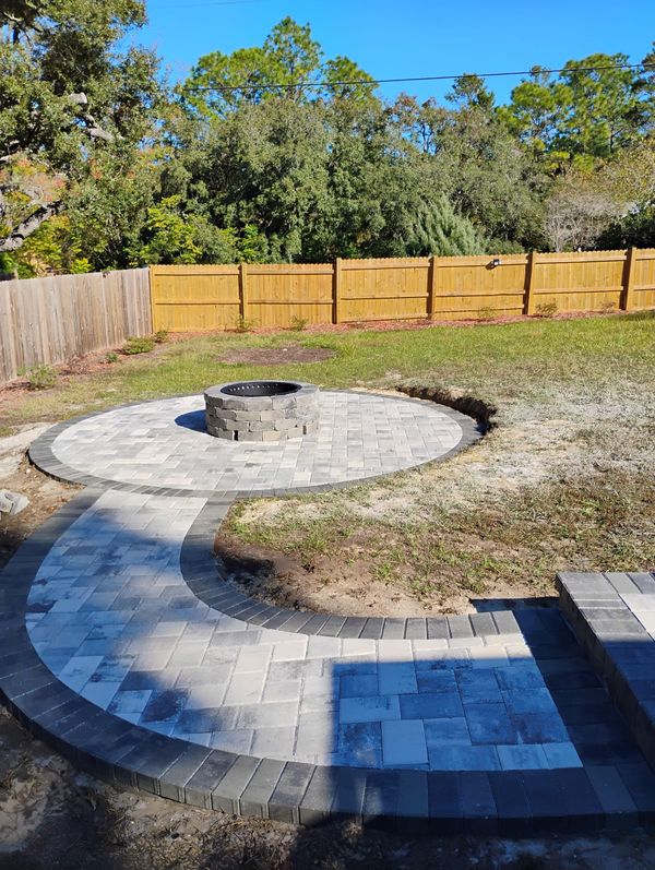 A stone fire pit sits in the center of a circular paver patio, connected by a curved walkway to an adjacent paved area.