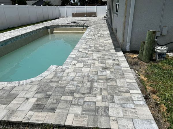 A rectangular swimming pool surrounded by a light-colored, multi-sized paver patio, with a white vinyl fence in the back.