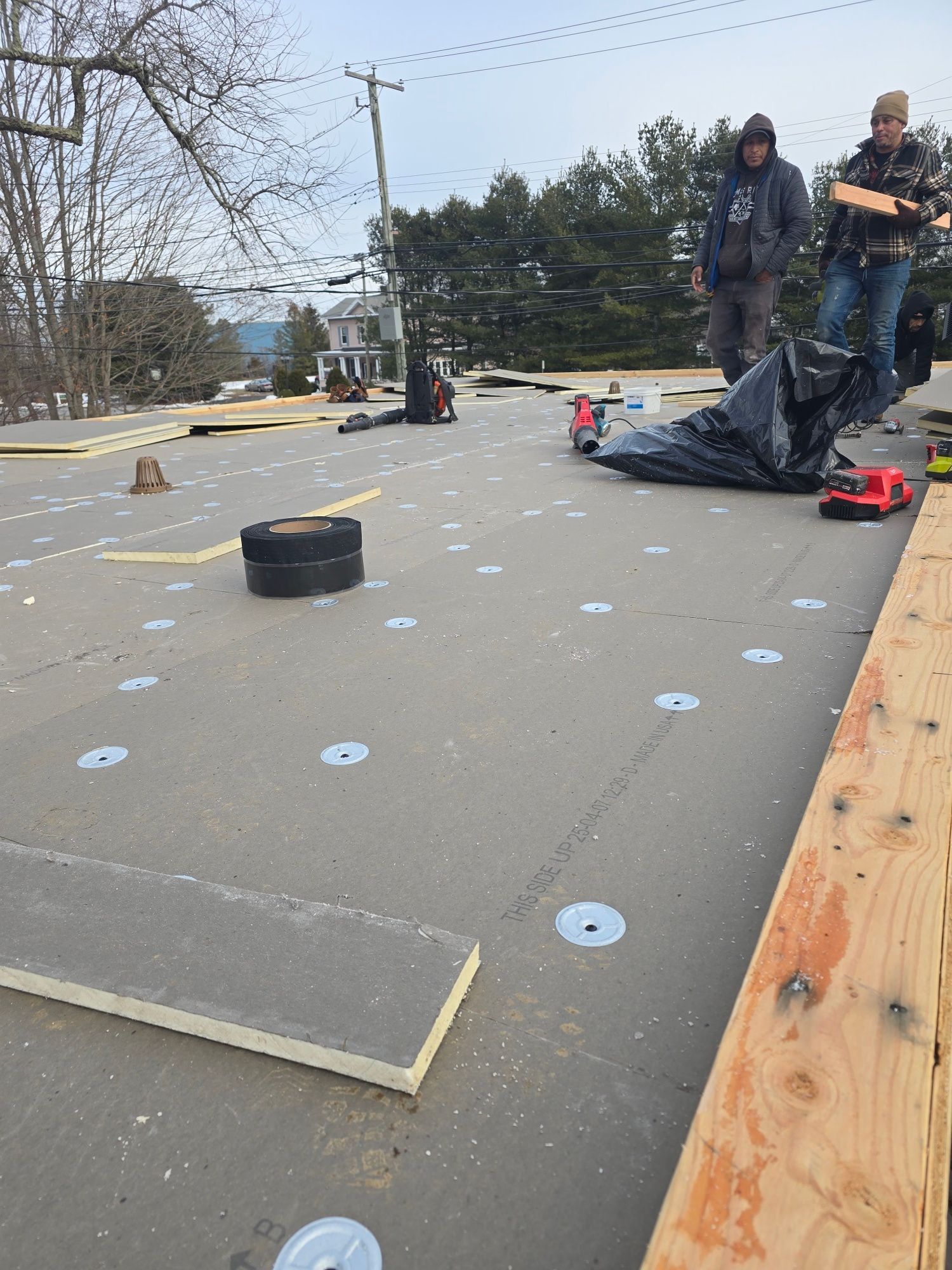 Roofing workers on a rooftop, laying down material. Gray material with silver discs, men holding tools, cloudy sky.